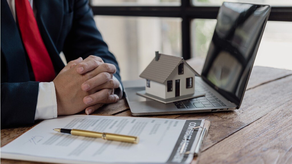 Mini house sitting on top of laptop with a pen and paper next to it and a person's hands showing