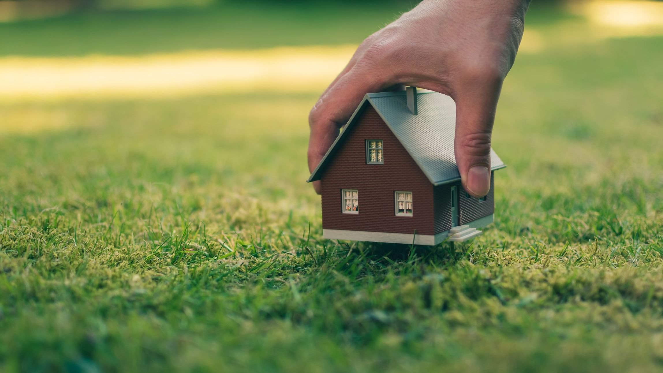 A hand is holding a model house above green meadow.