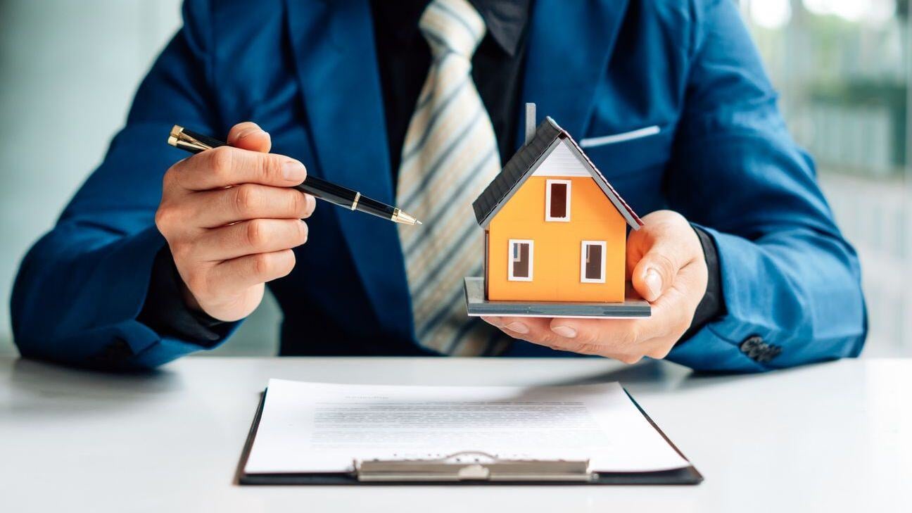 A person holding a pen and a small house, with papers on a desk.