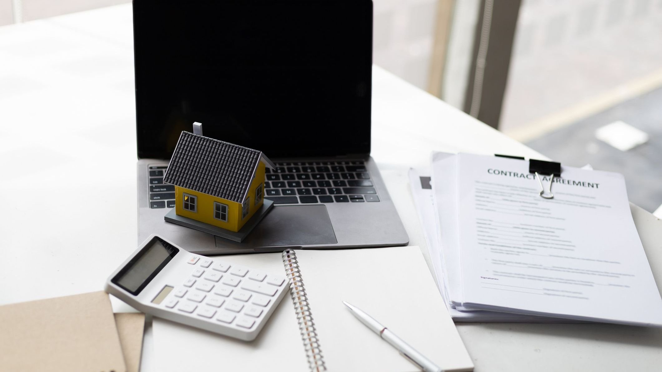 Wooden house, calculator, and stack of papers with a pen sitting on top.