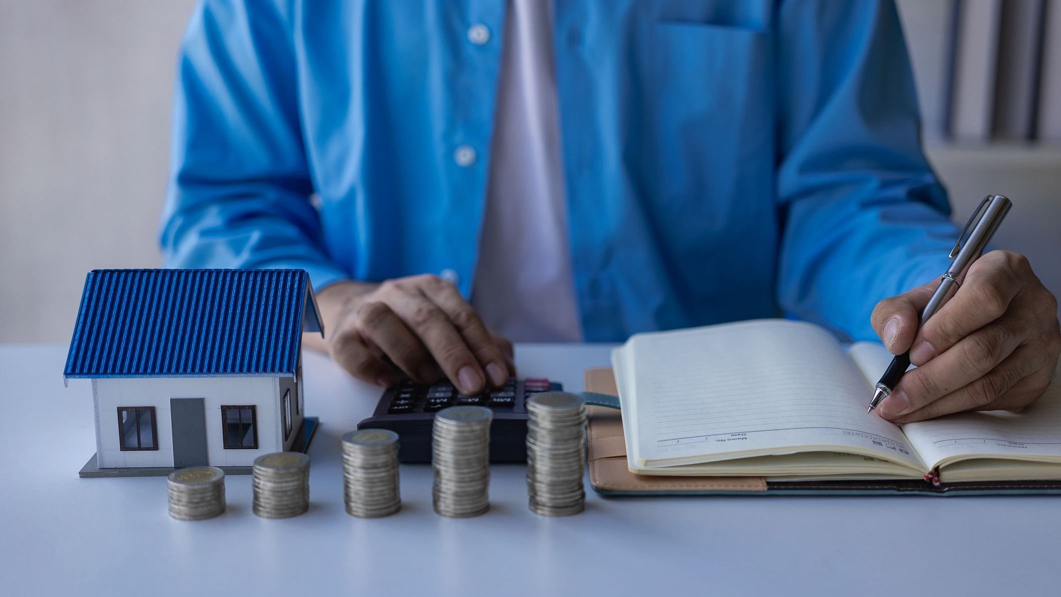 Man calculating notes on book and stack of coins lying on wooden table.