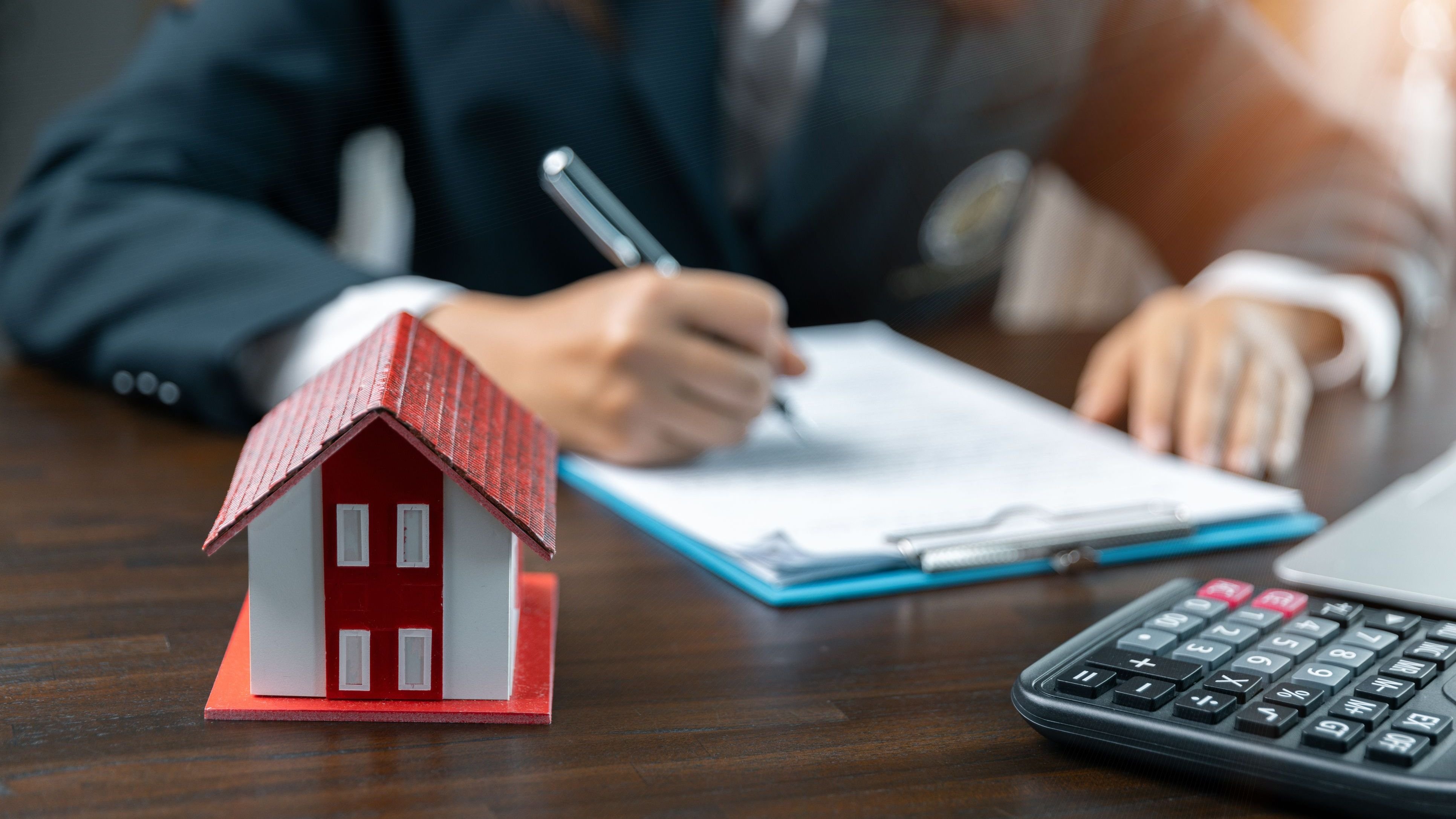 person signing papers with a house and calculator on the desk.