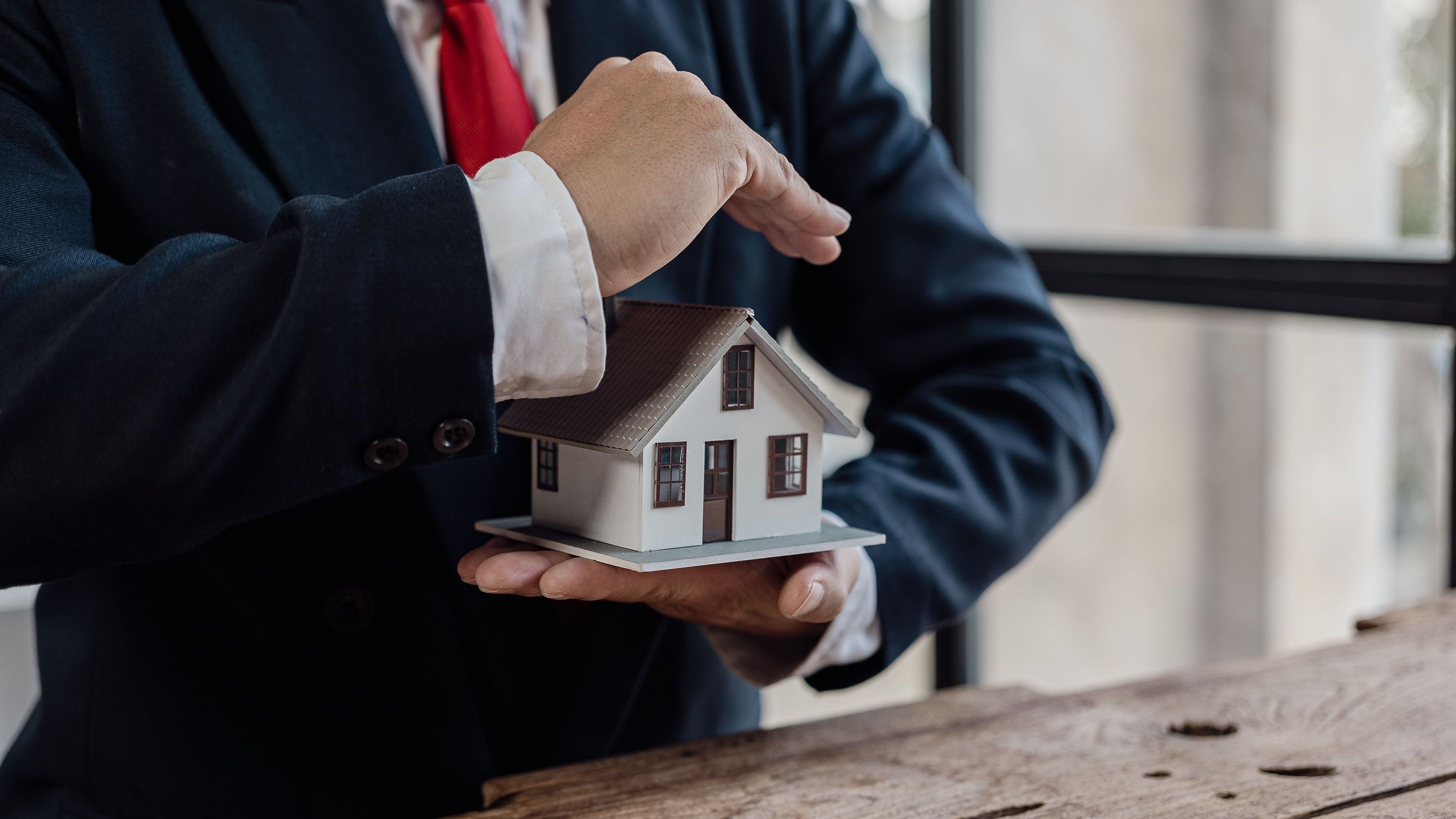 A businessman in a suit holds a house model in the palm of his hand.