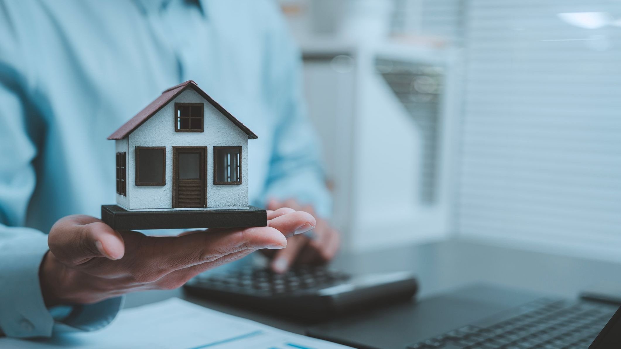 Real estate investment mortgage planning concept businessman holding miniature model house typing laptop computer keyboard analyzing housing market property valuation online