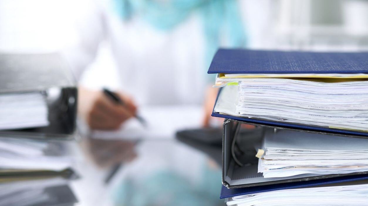 Person signing papers and a stack of papers sitting on desk.
