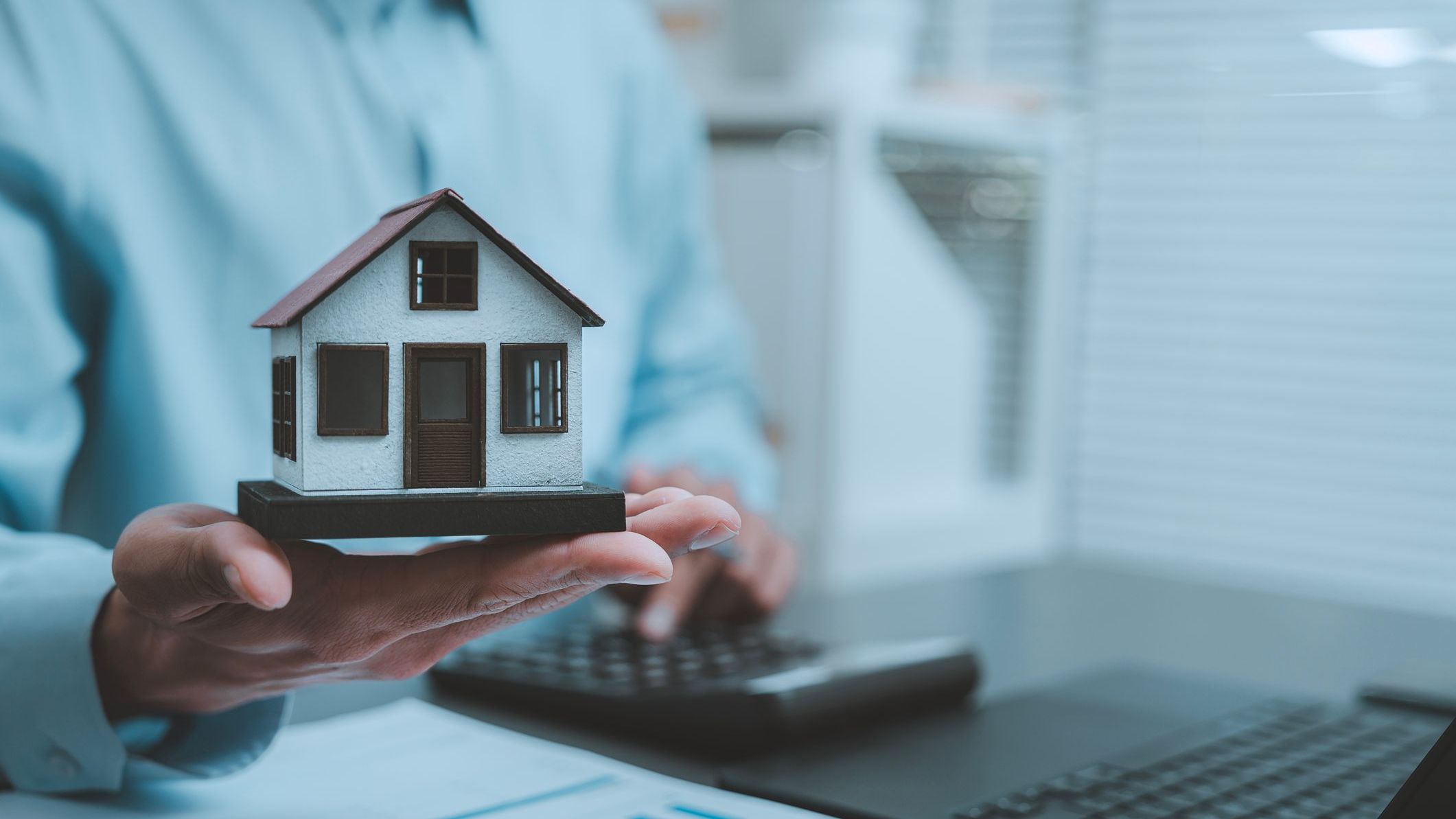 Real estate investment mortgage planning concept businessman holding miniature model house typing laptop computer keyboard analyzing housing market property valuation online