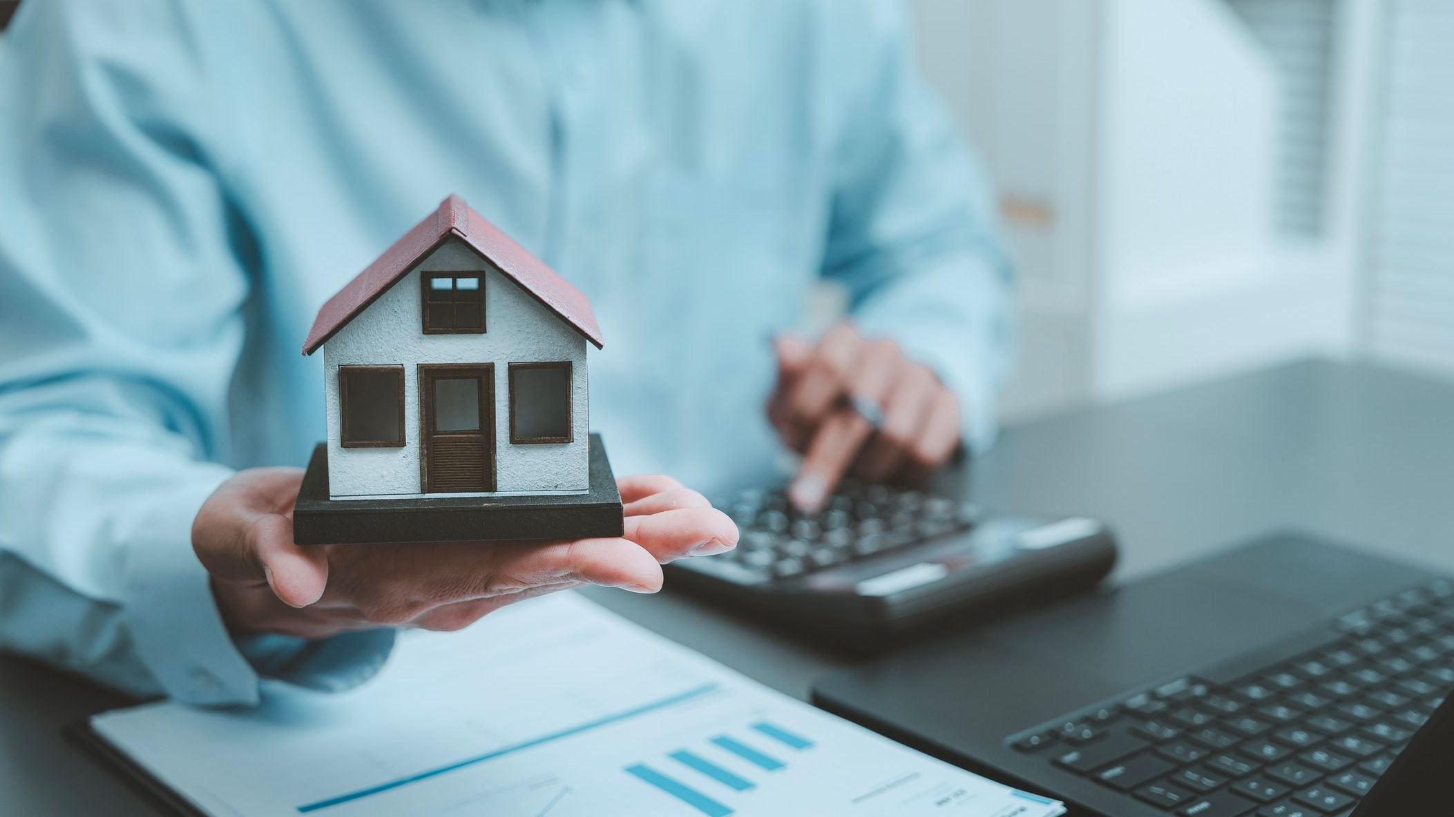 Real estate investment mortgage planning concept businessman holding miniature model house typing laptop computer keyboard analyzing housing market property valuation online